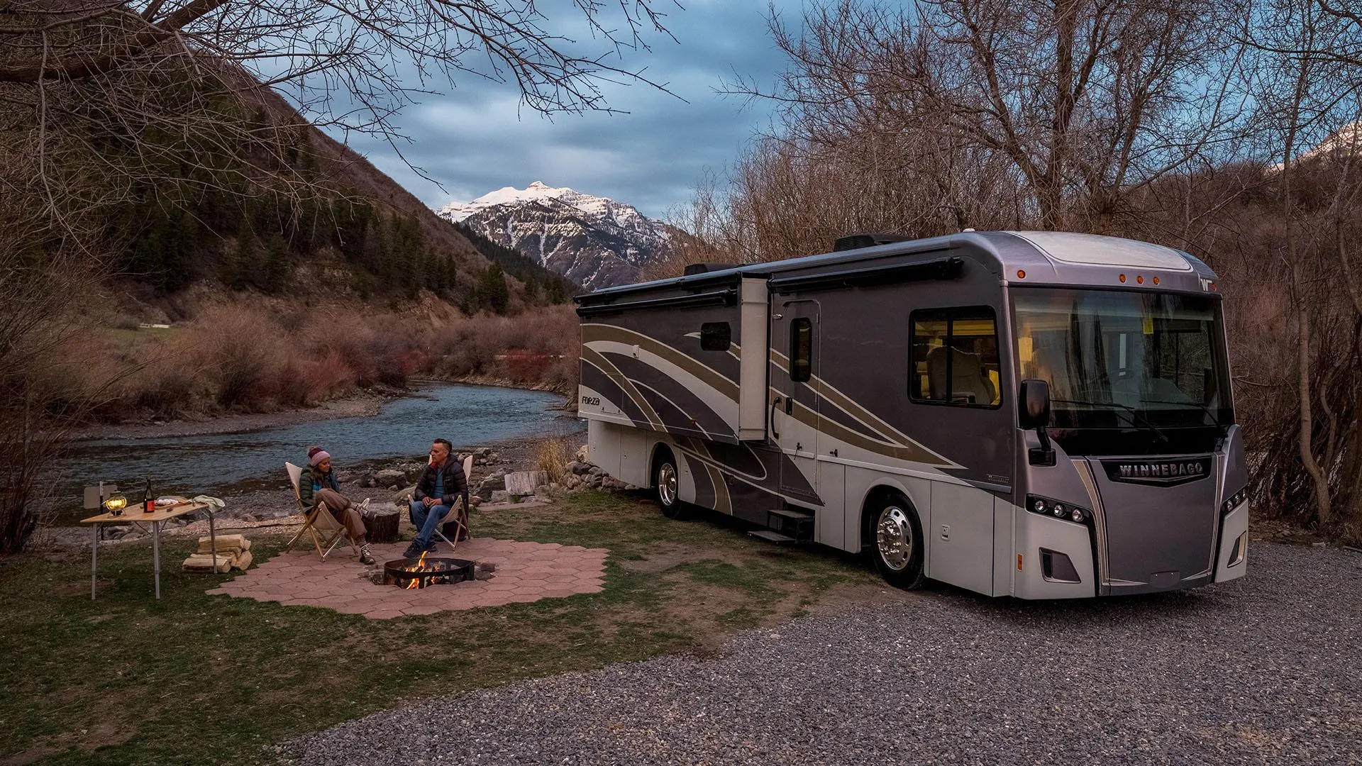 Winnebago motorhome parked by a river with campfire and snow-capped mountains